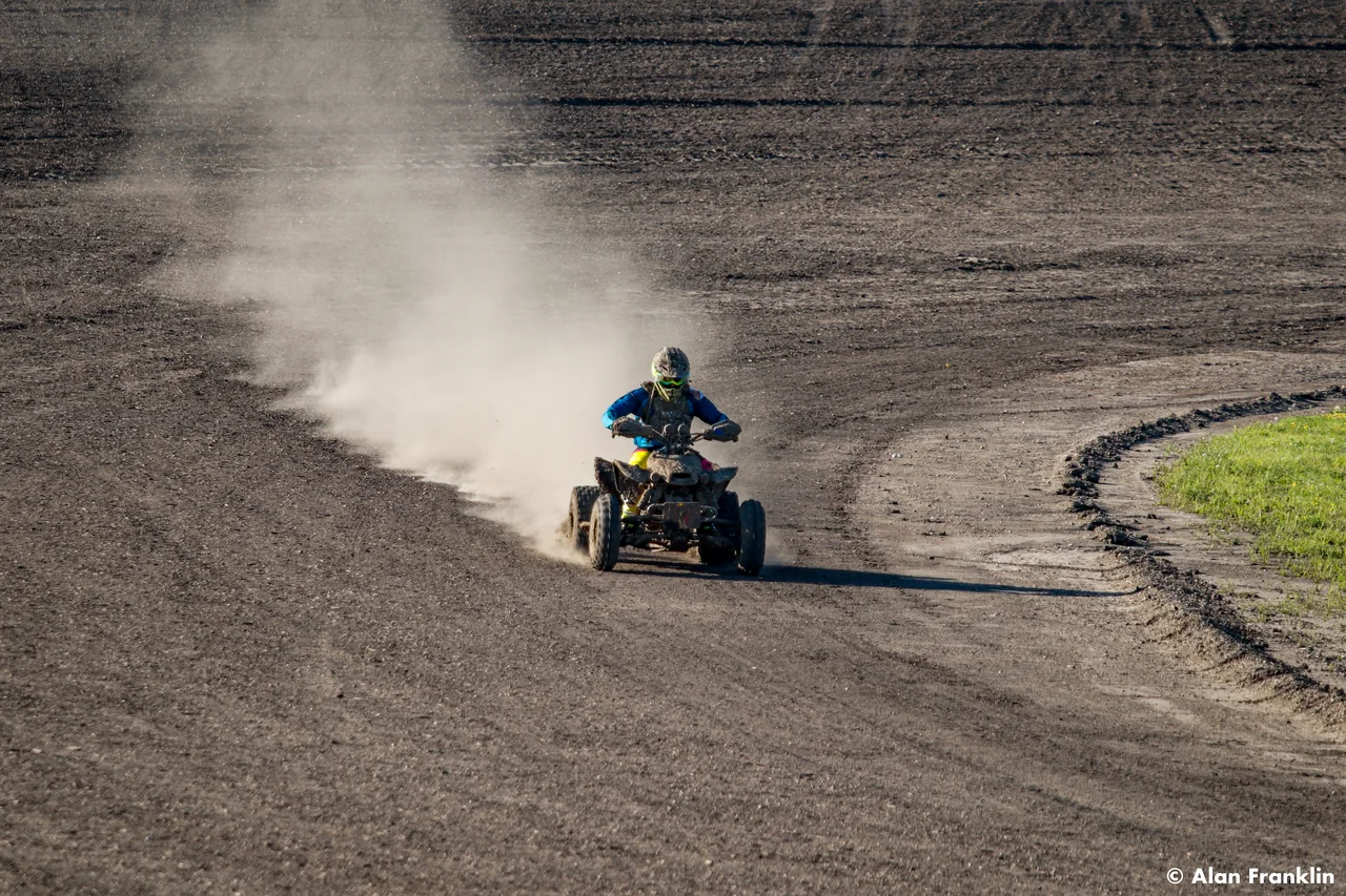 An ATV rider rounds a bend, trailing a long cloud of dust across the track.