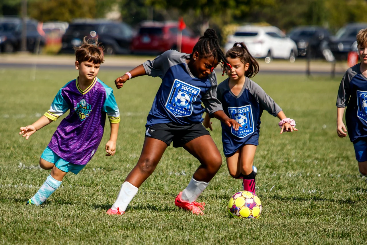 Three young soccer players compete for a yellow and purple ball in bright afternoon light.