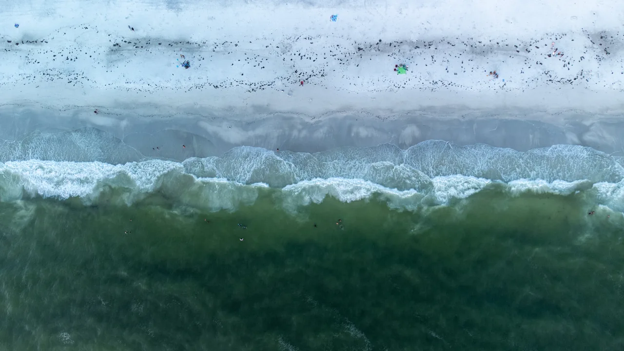 An overhead view of pale surf rolling onto a beach dotted with tiny figures.