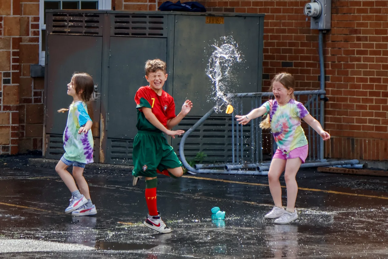 Children jump and laugh as a burst of water hangs in the air at a school field day.